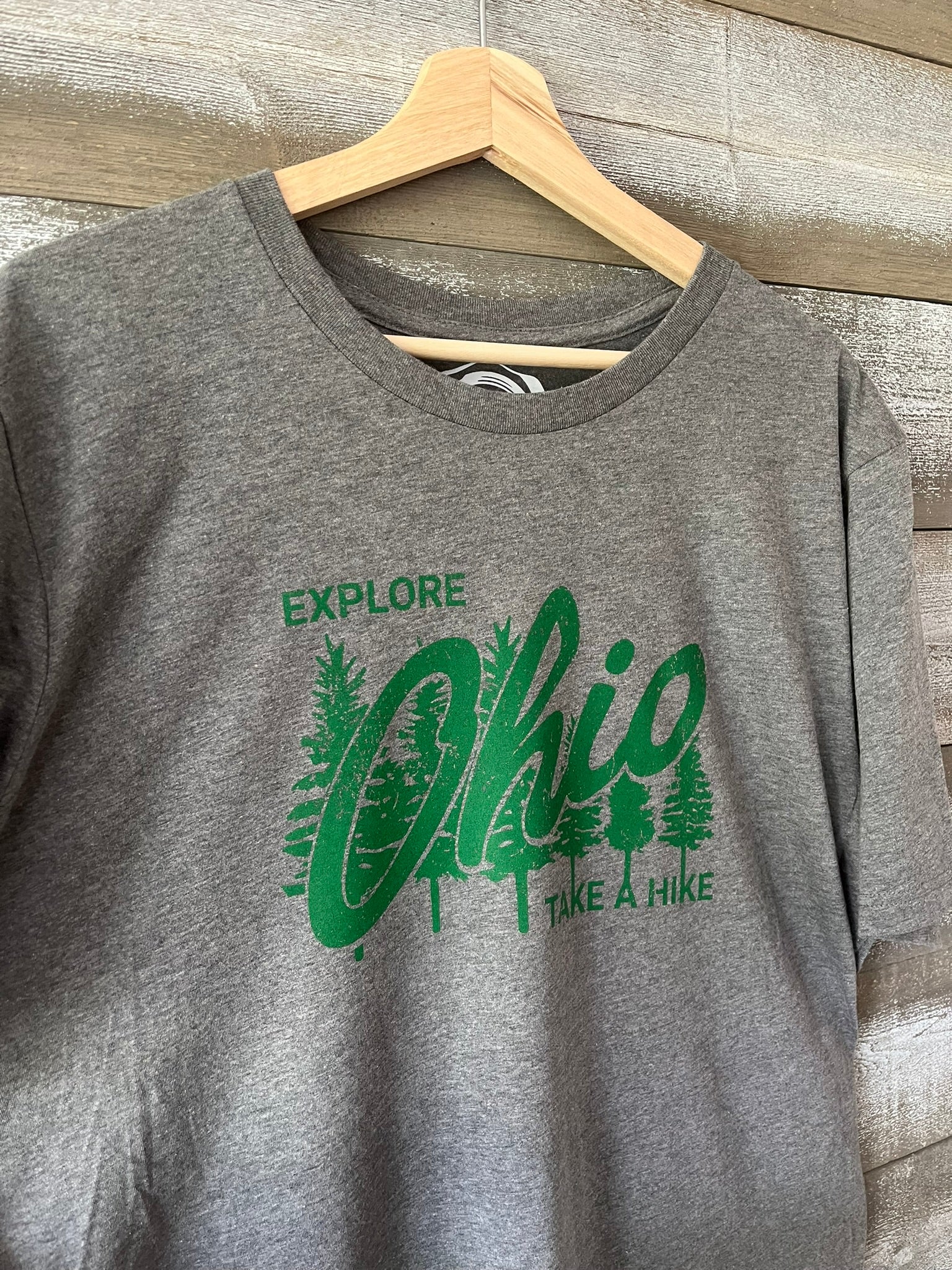 Gray t-shirt with green 'Explore Ohio Take a Hike' design on a wooden hanger.