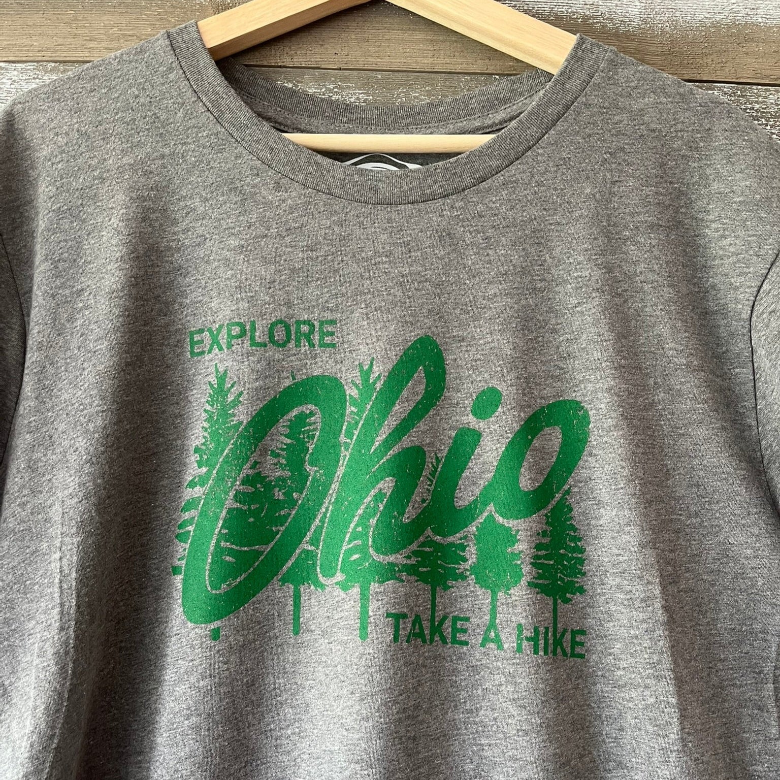 Gray t-shirt with green 'Explore Ohio, Take a Hike' text and graphics on a wooden hanger.